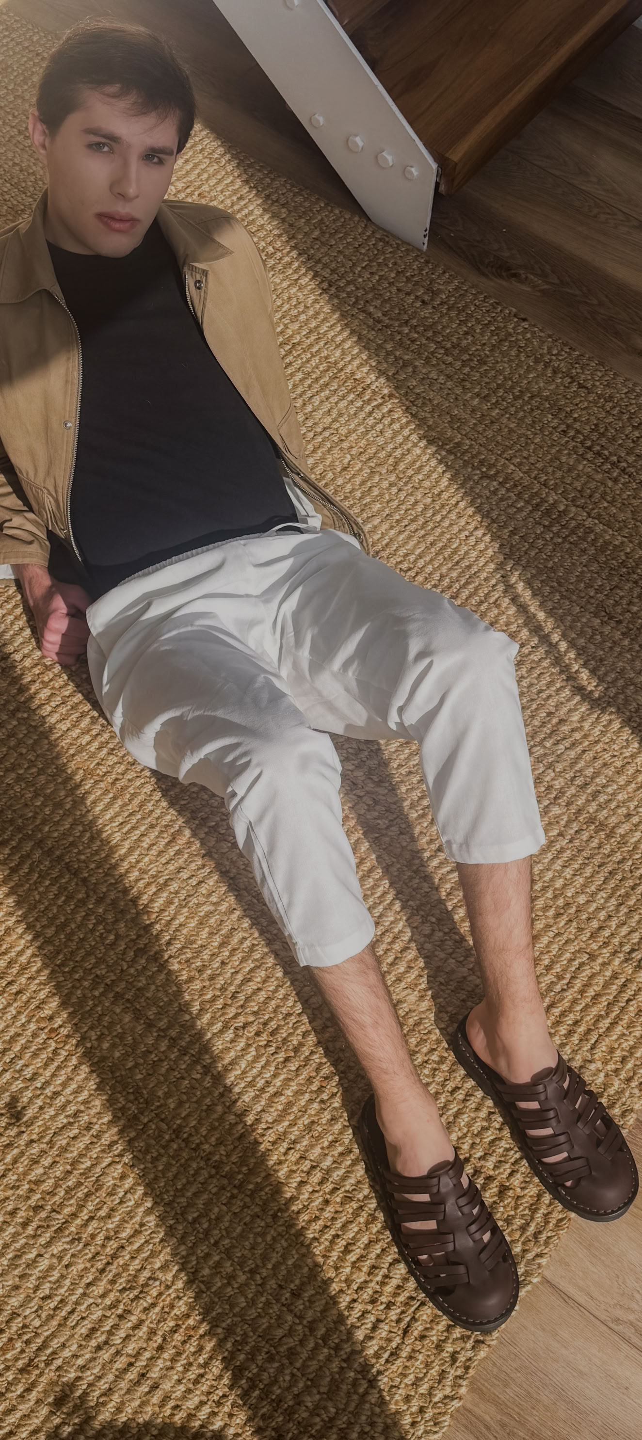 A fashionable man relaxes on a woven rug, wearing Debrovka handmade luxury leather sandals paired with white trousers, a black shirt, and a tan jacket. The image highlights the brand’s dedication to premium craftsmanship, comfort, and timeless style, making Debrovka sandals the perfect choice for modern men’s wardrobes.