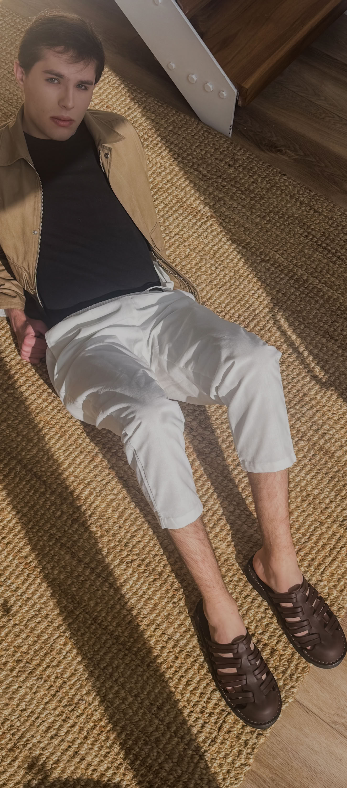 A fashionable man relaxes on a woven rug, wearing Debrovka handmade luxury leather sandals paired with white trousers, a black shirt, and a tan jacket. The image highlights the brand’s dedication to premium craftsmanship, comfort, and timeless style, making Debrovka sandals the perfect choice for modern men’s wardrobes.
