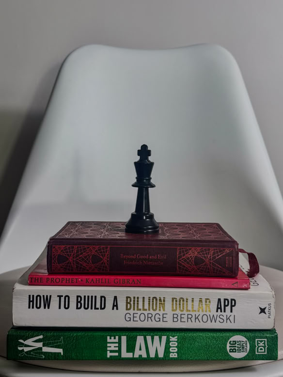 A striking composition featuring a stack of books on philosophy, business, and law, topped with a black chess king piece, set on a modern white chair. This image symbolizes the intelligence, strategy, and thoughtful craftsmanship that define Debrovka’s handmade luxury leather sandals and premium leather goods.