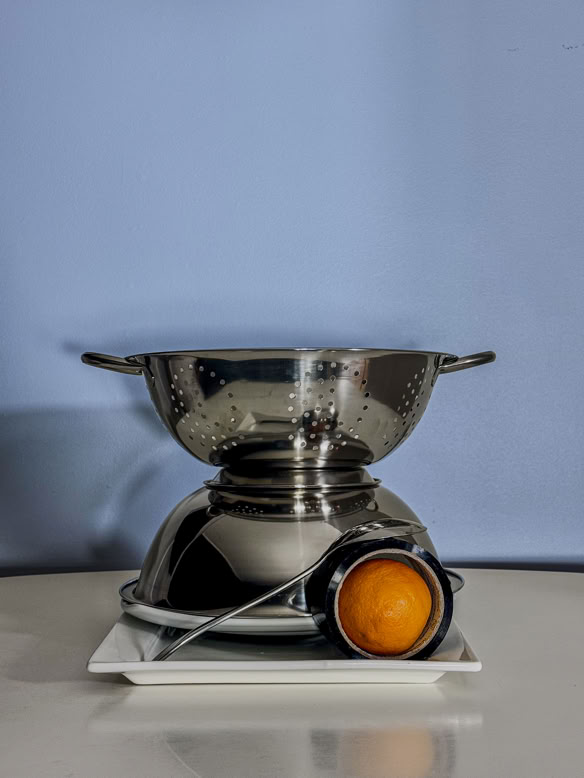 A modern still life featuring a stainless steel colander, bowl, and a single orange in a metal scoop, arranged on a white plate against a soft blue background. This image reflects Debrovka’s appreciation for everyday artistry and meticulous craftsmanship, the same qualities found in every pair of their handmade luxury leather sandals and premium leather goods.